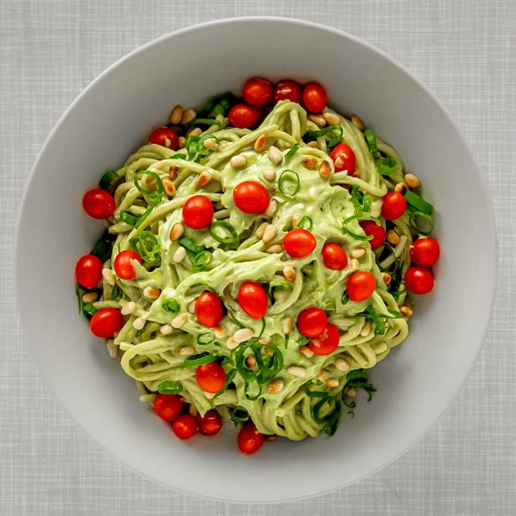 Simple and creamy avodado pasta, seen from top, topped with pine nuts, cocktail tomatoes and spring onions