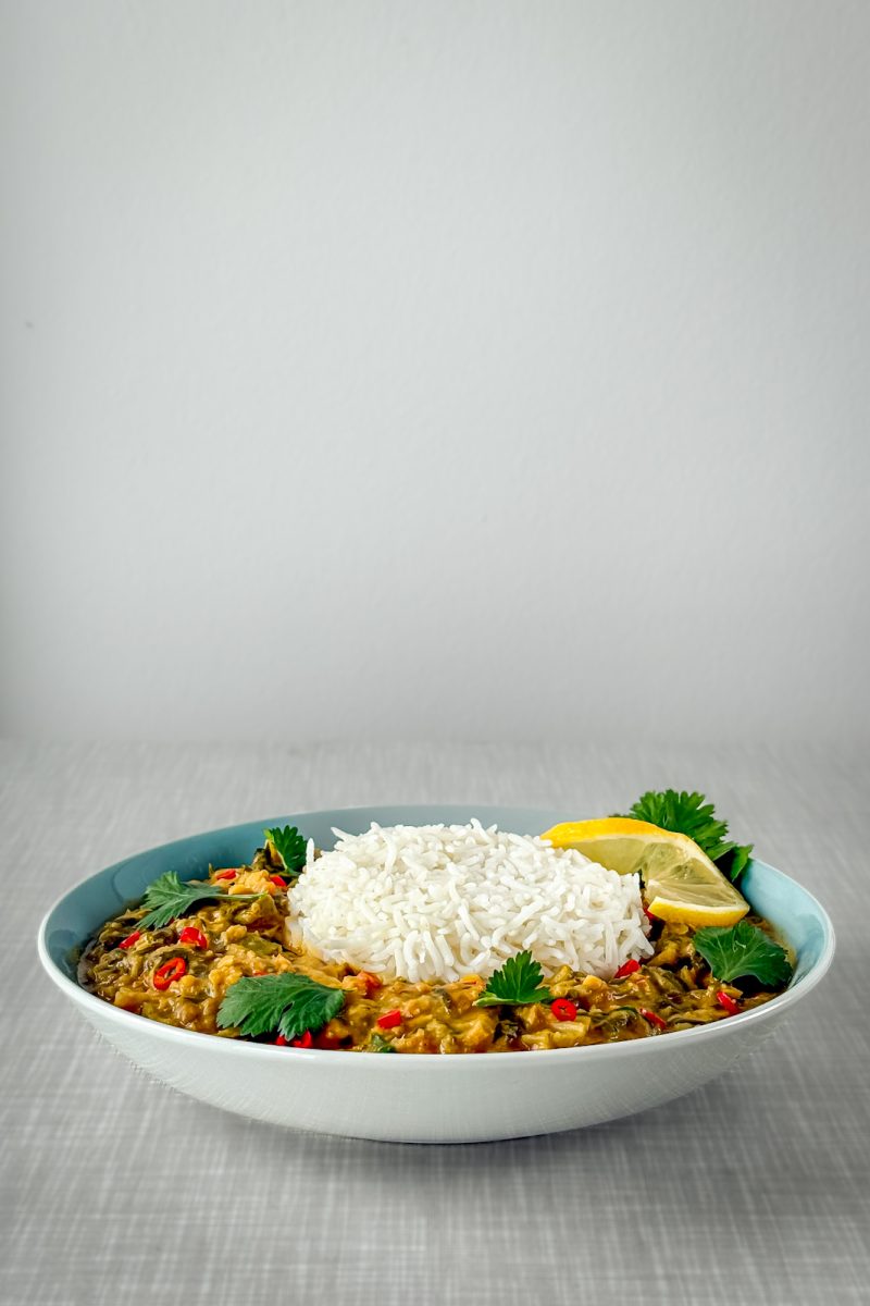 A turquoise plate featuring a creamy Swiss chard and lentil curry served with basmati rice, garnished with cilantro leaves, a twisted slice of lemon, and chili rings. Photographed from a diagonal front angle.