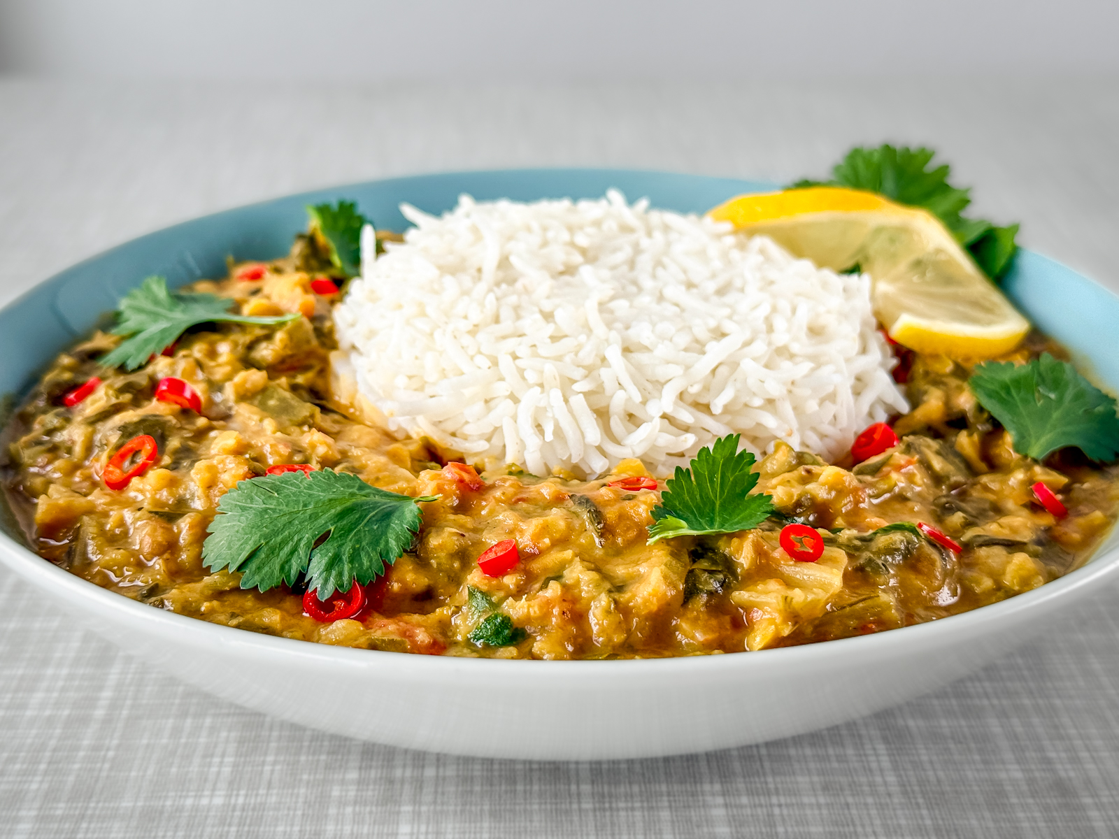 Close-up of a plate of Swiss chard and lentil curry with rice. Garnished with fresh cilantro, a slice of lemon, and chili rings. Photographed from a diagonal front angle.