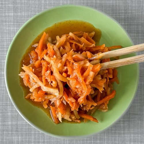 Ein kleines pastellgrünes Tellerchen, gefüllt mit einem Karotten Kohlrabi Radieschen-Kimchi, von oben fotografiert. Zwei Essstäbchen halten einen Happen des Kimchi.
