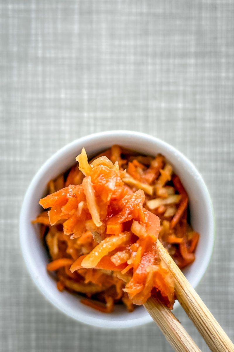 Zwei Essstäbchen halten eine Portion Karotten-Kimchi. Im Hintergrund die kleine Schüssel. Auf einem grauen Tisch. Von oben fotografiert.