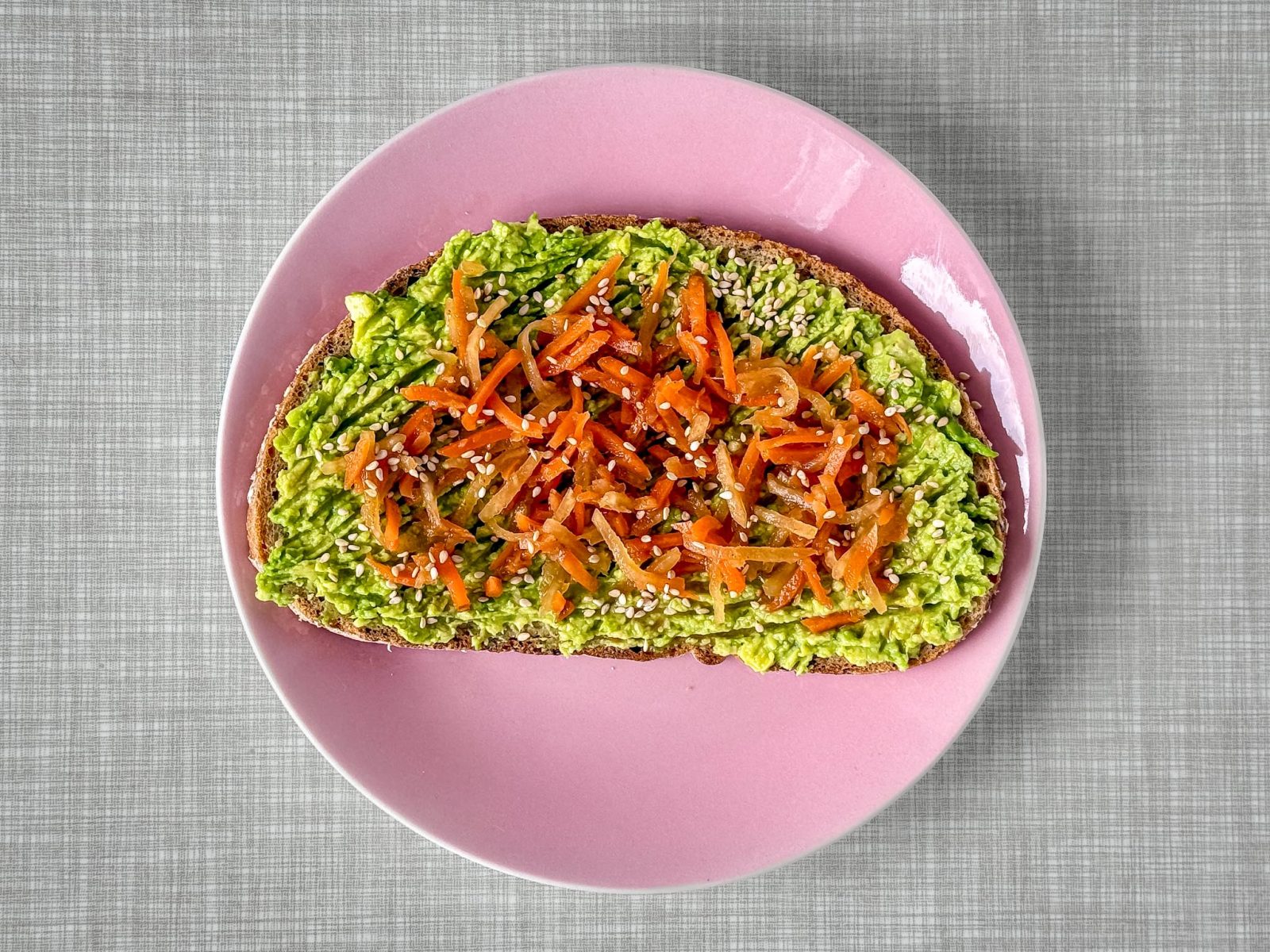 Ein pastellrosa Teller mit einem Avocado-Brot, getoppt mit einem Karotten Kimchi und Sesam.