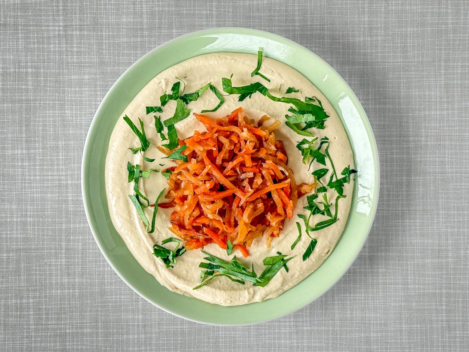 A pastel green bowl filled with hummus, topped with carrot kimchi and fresh parsley.