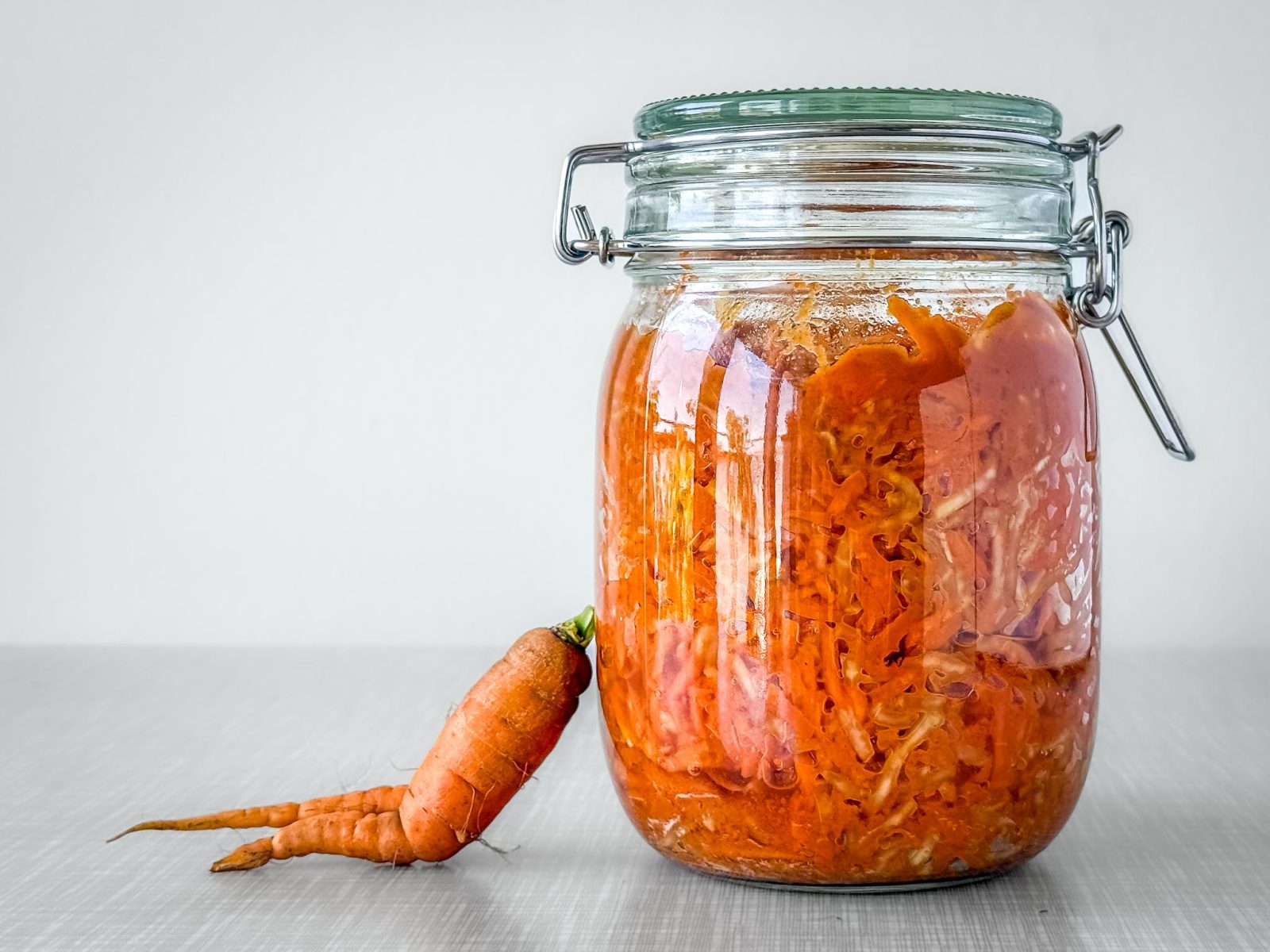 Ein Bügelverschluss-Glas gefüllt mit einem Karotten-Kohlrabi-Radieschen-Ferment. Am Glas lehnt sich eine "zweibeinige" Karotte "sitzend" an.