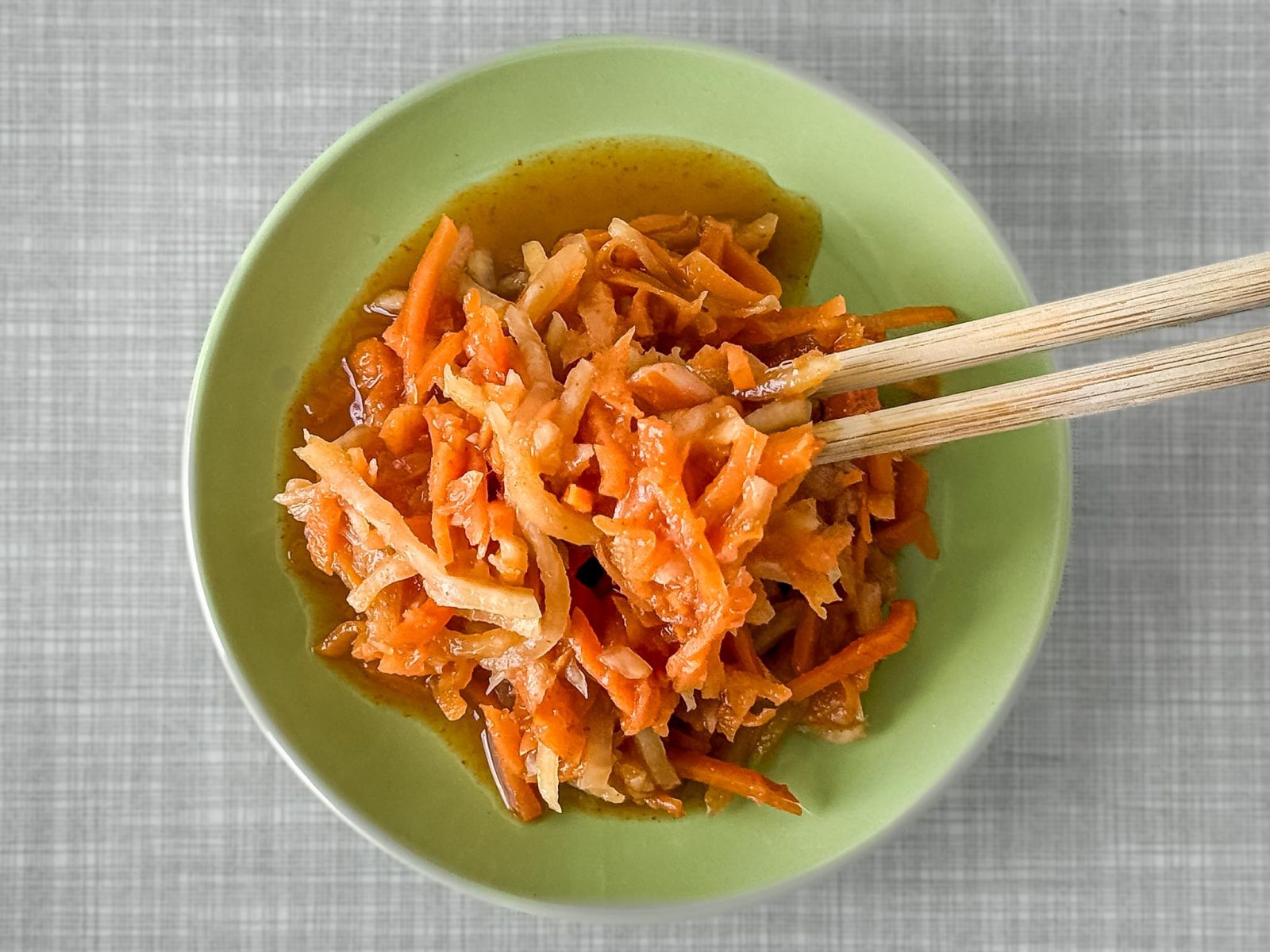 A small pastel-green plate filled with carrot, kohlrabi, and radish kimchi, photographed from above. Two chopsticks are holding a bite-sized piece of the kimchi.